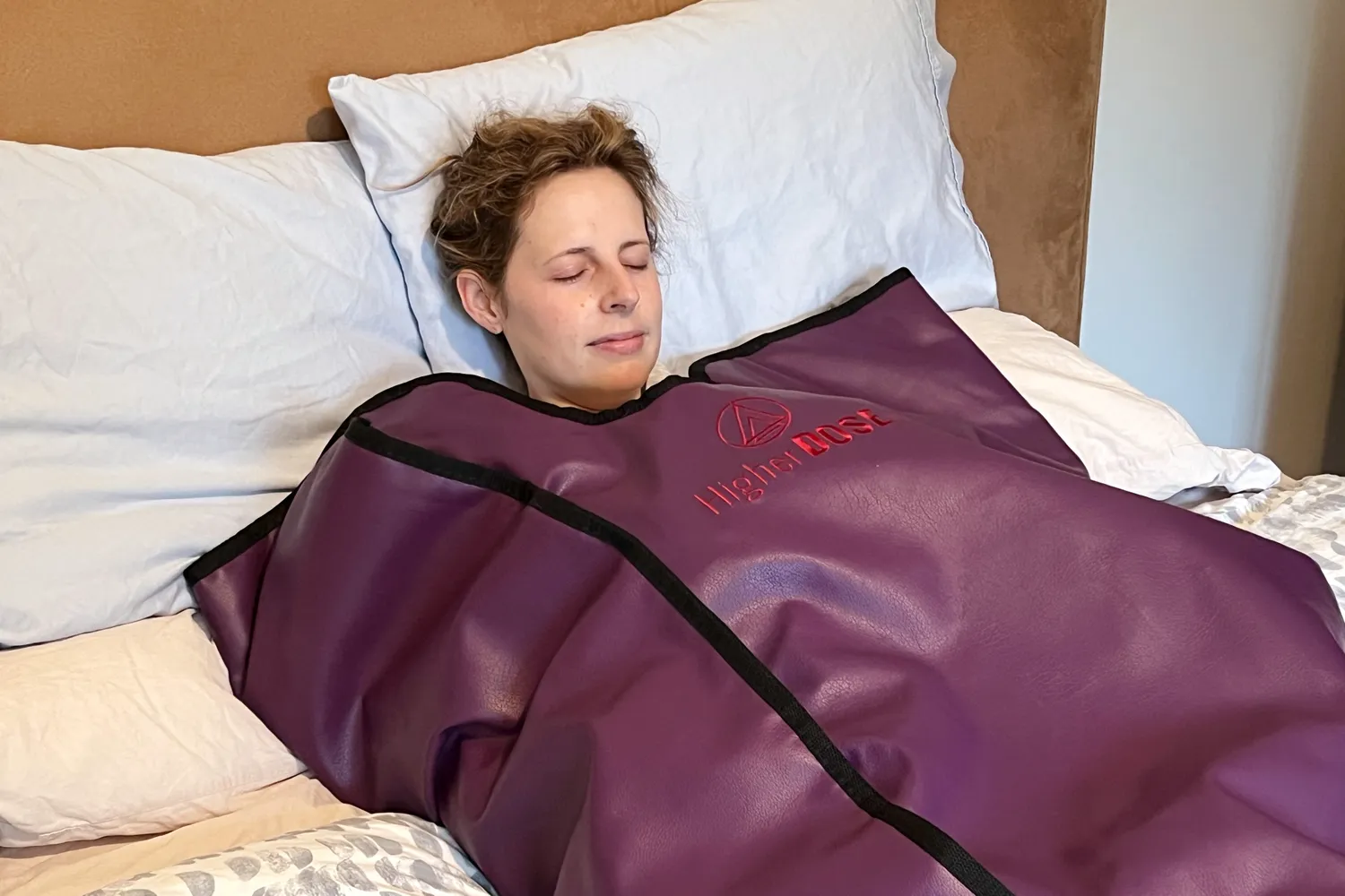 A person lying on a bed wrapped in an infrared sauna blanket with the Higher Dose logo visible