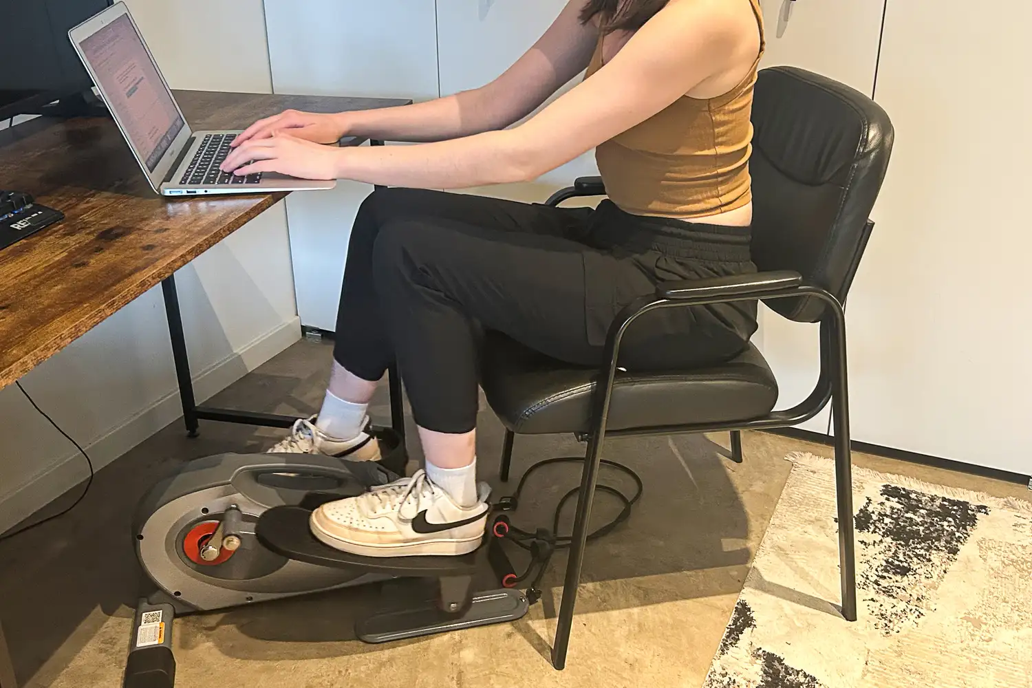 A person sits in a chair while using the Sunny Health & Fitness Smart Stand/Sit Mini Elliptical Exercise Trainer