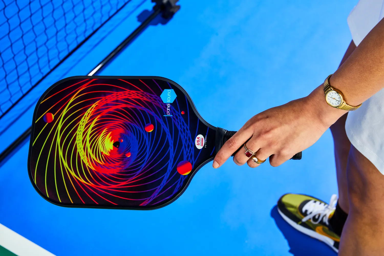 A hand holding a pickleball paddle with a vibrant spiral design on a blue court wearing a watch and rings sneaker and net partially visible